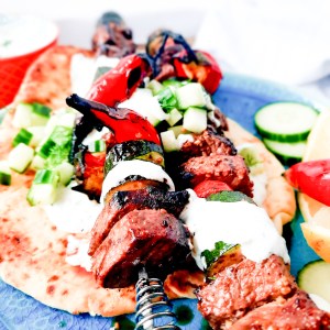 Tender, marinated Greek Inspired Steak Kabobs with Tzatziki, zucchini, mushroom, chili peppers and cucumber dill relish top pita bread.