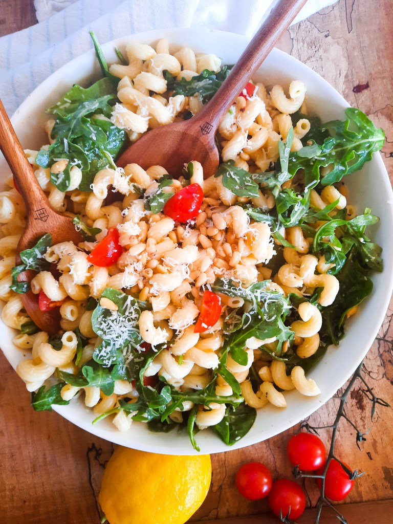 A white bowl filled with pasta, arugula, cherry tomatoes, and pine nuts, garnished with grated cheese. Two wooden serving spoons are placed in the bowl, and a lemon and some cherry tomatoes are positioned on the side.