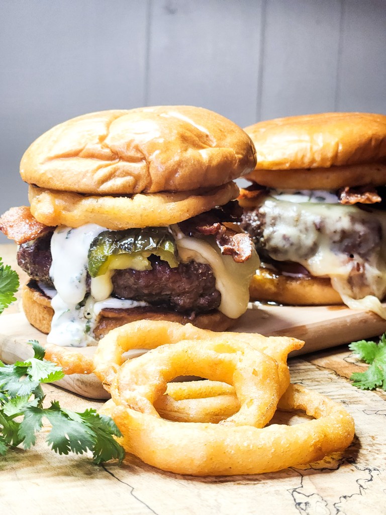 OUT WEST Roasted Jalapeno Burger piled high with bacon, onion rings, cilantro lime crema, creamy jack cheese and a buttery brioche bun.