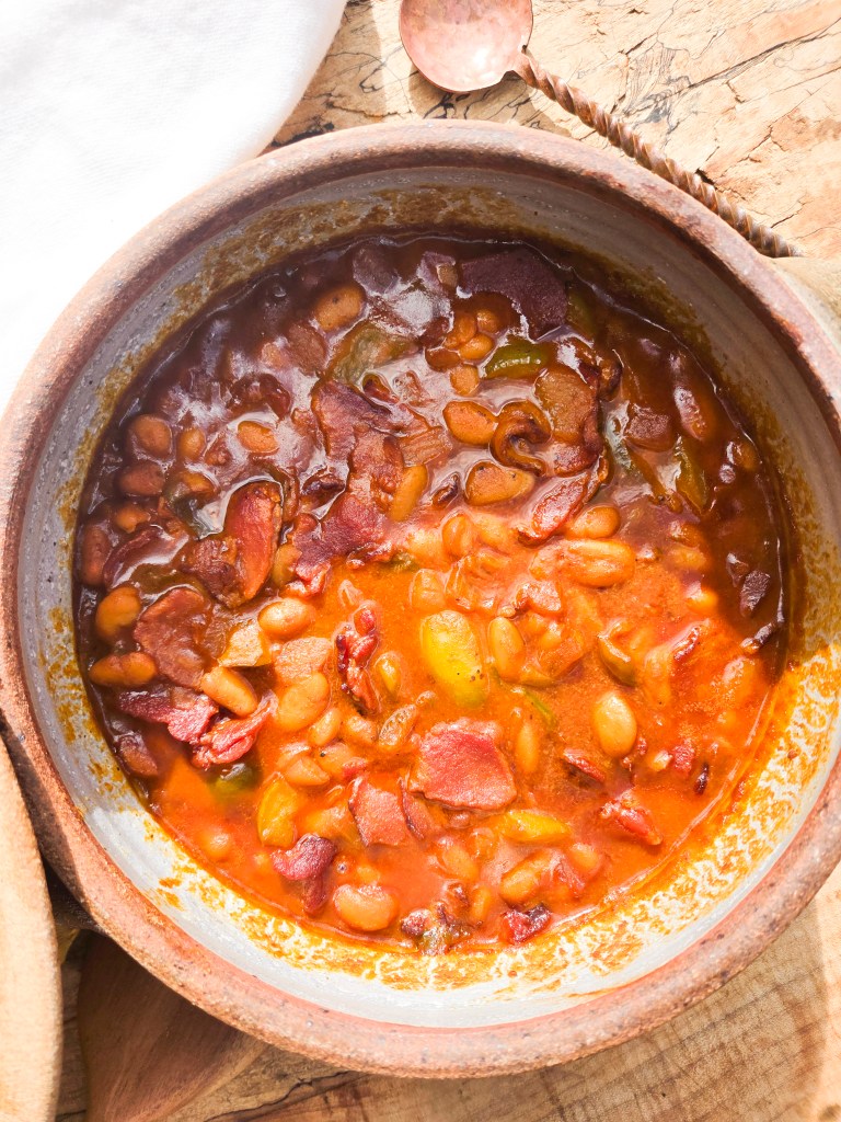 Close-up of a rustic clay pot filled with bean stew, featuring a rich tomato-based broth, pieces of bacon, and a variety of beans and vegetables.