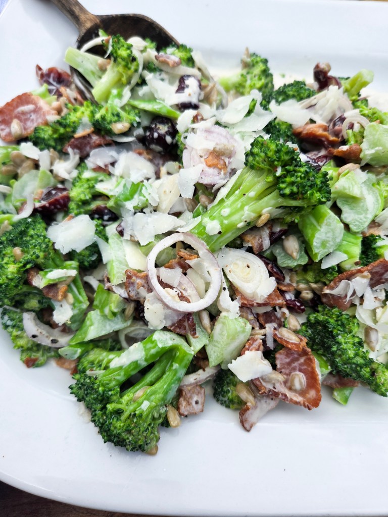 A close-up view of a fresh broccoli salad with ingredients like sliced onions, bacon, sunflower seeds, and grated cheese on a white plate.