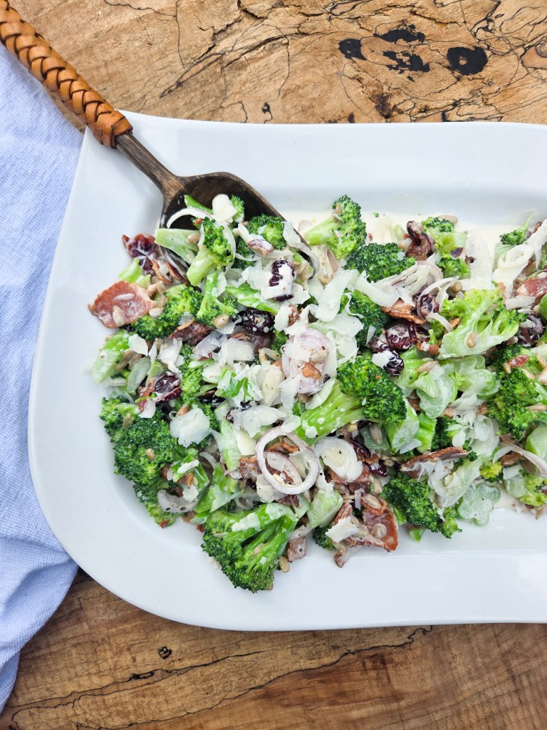 A fresh broccoli salad topped with sliced shallots, nuts, and a creamy dressing, served on a white platter against a wooden background.